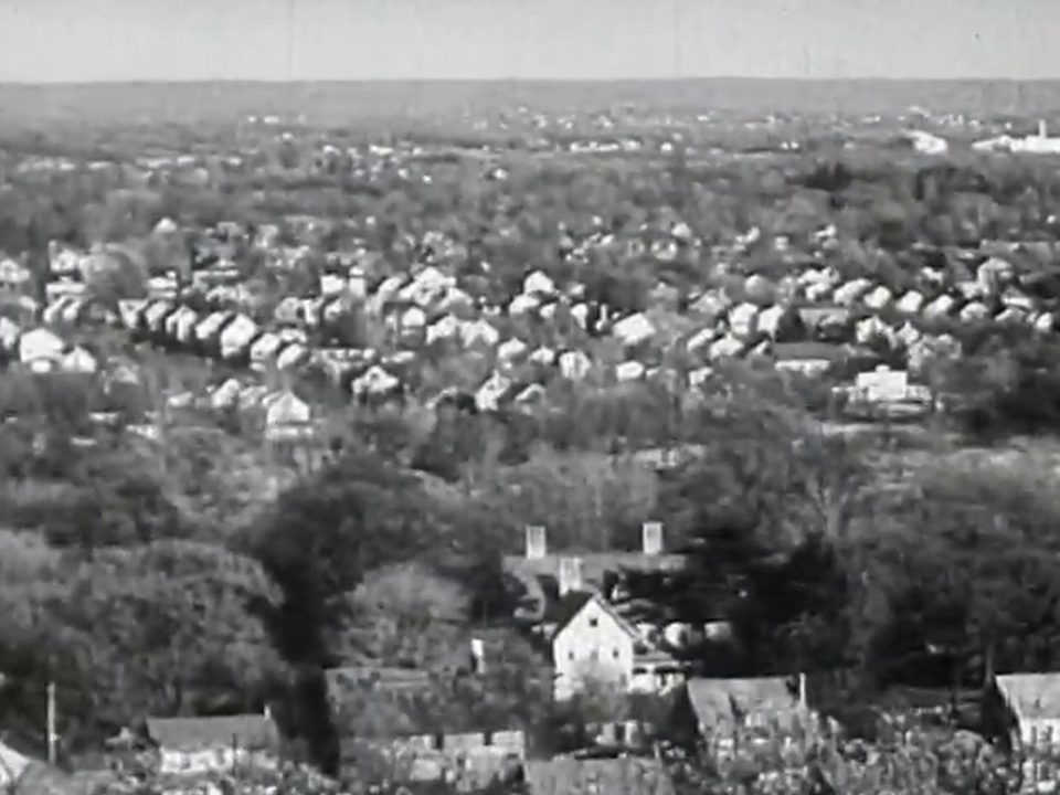 Black-and-white film still: a wide view from a hill over the rooftops and trees of a small town.